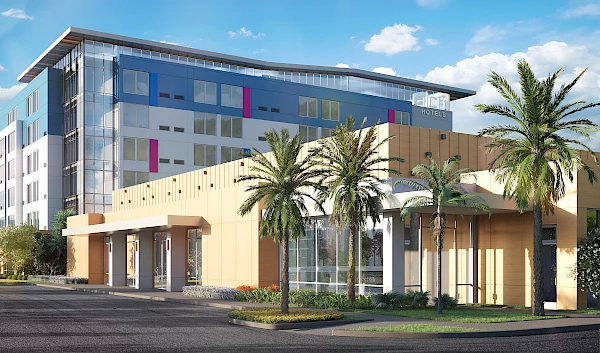 The image shows a modern hotel building with palm trees, featuring a glass and beige facade with colorful accents and a clear blue sky above it.