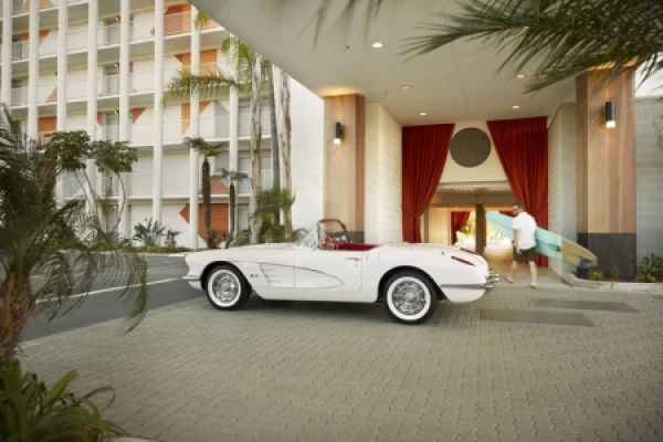 A white convertible car is parked under a building's entryway. A man holding a surfboard walks toward the entrance, surrounded by palm trees.
