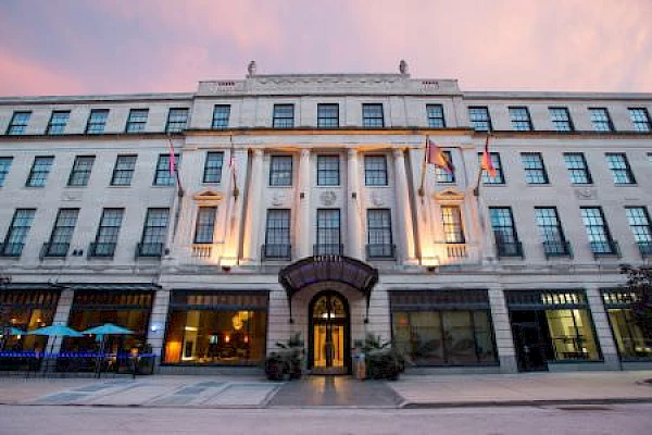 A grand, multi-story building with columns, flags, and large windows is shown at dusk, illuminated by ambient lights against a pink sky.