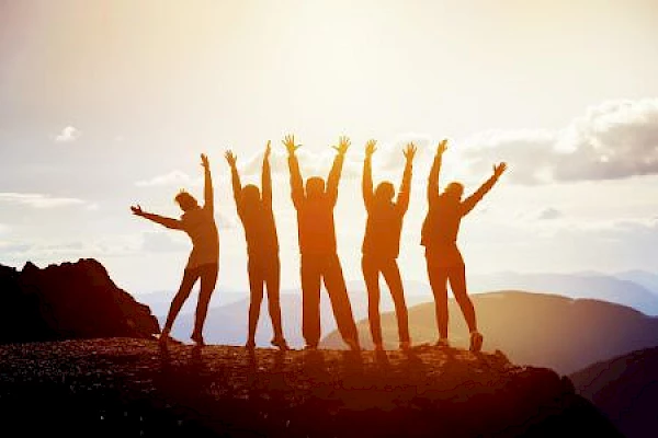 A group of five people stand silhouetted on a mountain top with arms raised in celebration, backlit by the setting sun.