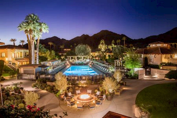 A luxurious resort with a pool, firepit seating area, lit-up palm trees, and mountainous backdrop under an evening sky, creating a serene atmosphere.