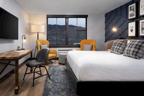 A cozy, modern hotel room with a bed, desk, chairs, and wall art. A window offers a view of mountains in the distance, enhancing the decor.