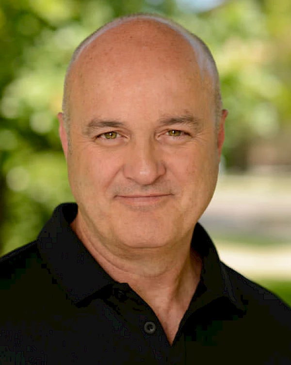 A man with a shaved head wearing a black shirt smiles at the camera, with a blurred green outdoor background.