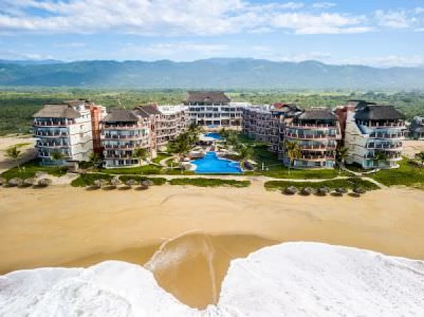 Aerial view of a beach resort with multiple buildings, a central pool, green area, and mountains in the background, right by the ocean.