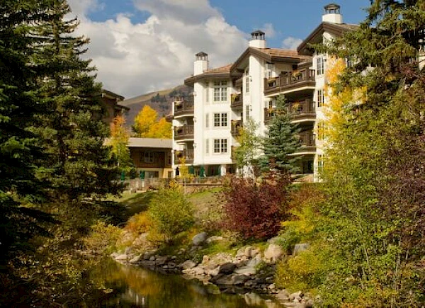 An elegant multi-story building is nestled among dense trees with a clear stream flowing in the foreground and mountains in the background.