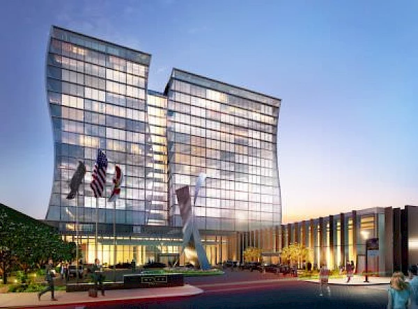 The image shows a modern, curvy glass building with flags in front, a large sculpture, and people walking around the entrance during sunset.