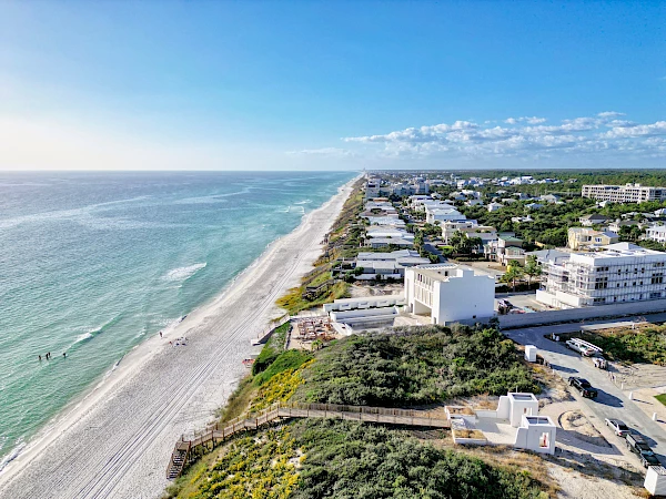 An aerial view of a coastal town featuring a beach, clear blue water, residential buildings along the shoreline, and a calm, sunny day.