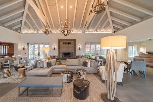 A cozy living room with vaulted ceilings, modern furniture, two chandeliers, floor lamps, and a fireplace in the background, creating a warm atmosphere.