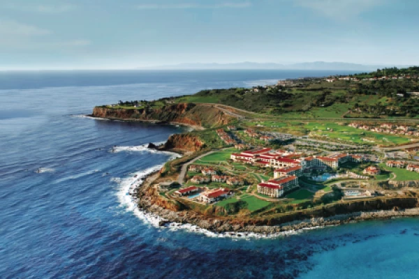 A coastal resort with red-roofed buildings sits on a cliff overlooking the ocean, surrounded by lush greenery and cliffs.