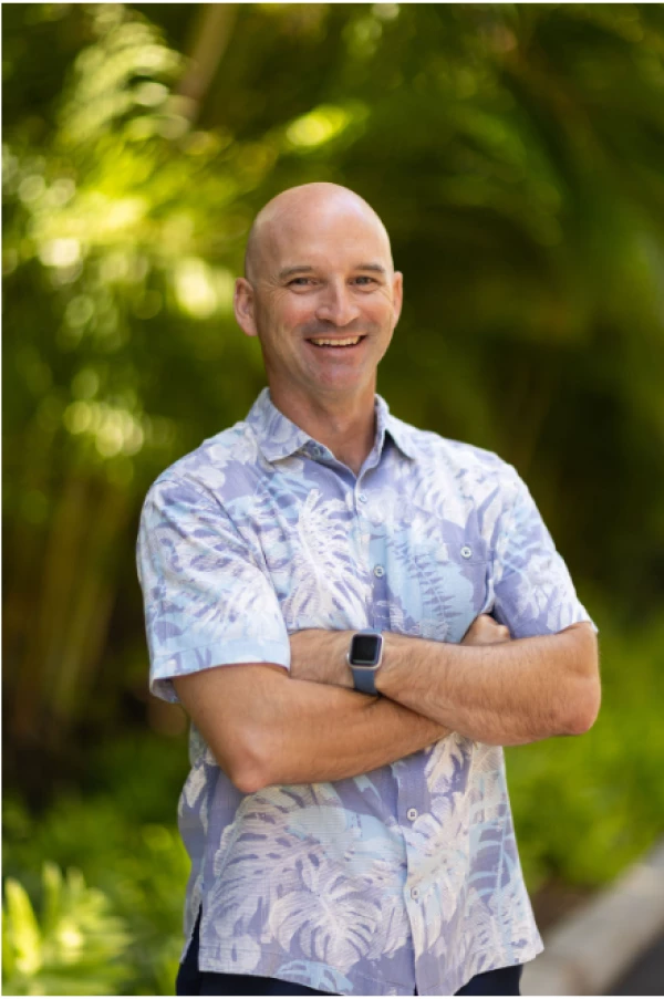 A person wearing a floral shirt and a smartwatch stands outdoors with arms crossed, smiling, and green foliage in the background.