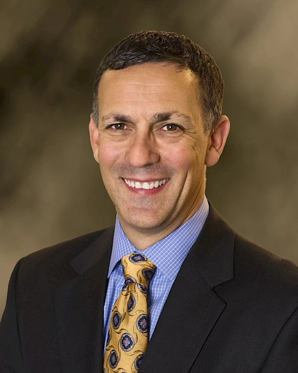 A man in a suit and patterned tie smiles at the camera against a blurred background.