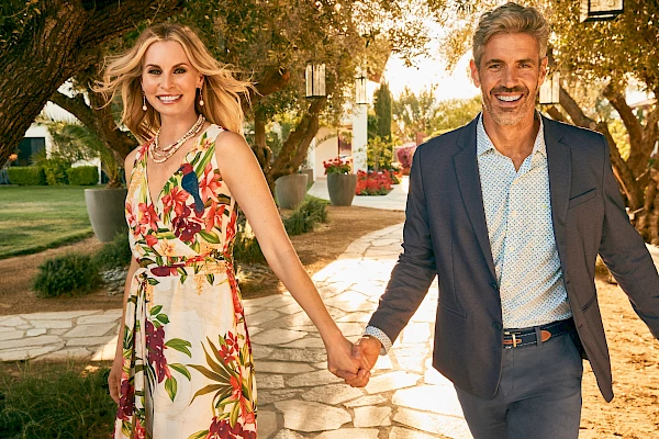 A smiling couple is holding hands, walking down a stone path in a garden with trees and potted plants, dressed in casual yet elegant attire.