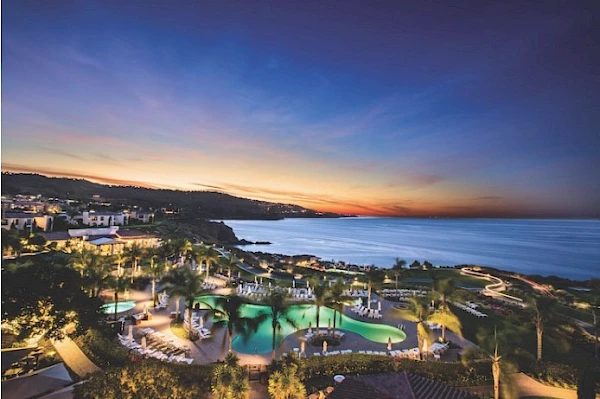 This image showcases a luxurious coastal resort at sunset, featuring pools and lush greenery overlooking a serene ocean view.