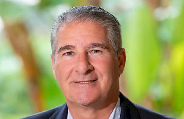 A man with short gray hair smiling, wearing a dark suit, with a blurred green leafy background.