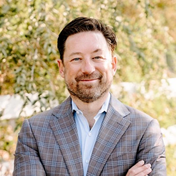 The image shows a man with short hair and a beard, wearing a checked blazer and a light blue shirt, standing outdoors with folded arms.