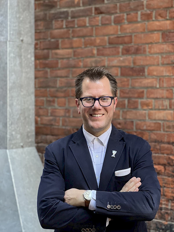 A man in a suit stands against a brick wall with arms crossed, wearing glasses and smiling.