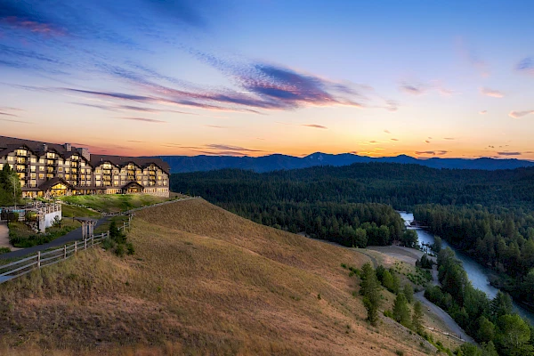 A scenic view of a lodge on a hillside during sunset, overlooking a lush forested valley with a winding river, under a sky with colorful clouds.