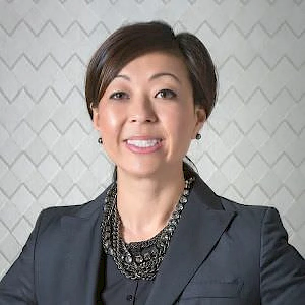 A person with short hair is smiling, wearing a dark blazer and a layered necklace, against a patterned background.