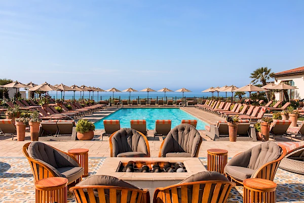 An outdoor pool area with lounge chairs, umbrellas, and cushioned seating around a fire pit, overlooking the ocean under a clear blue sky.