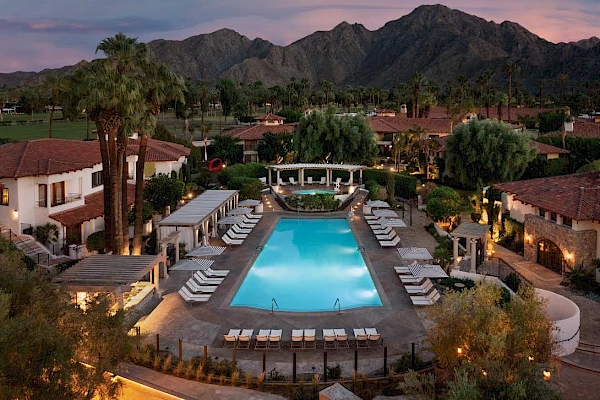 A resort features a central swimming pool with numerous lounge chairs, surrounded by buildings, palm trees, and mountains in the background.